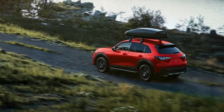 2027 Honda HR-V Sport in Milano Red profile view driving on a dirt road near water with a roof box (Subcompact Crossover SUV).